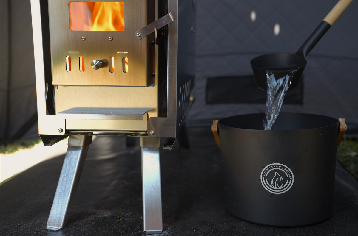 Bamboo handled ladle pouring water into a black aluminum sauna bucket next to a wood stove in a tent sauna