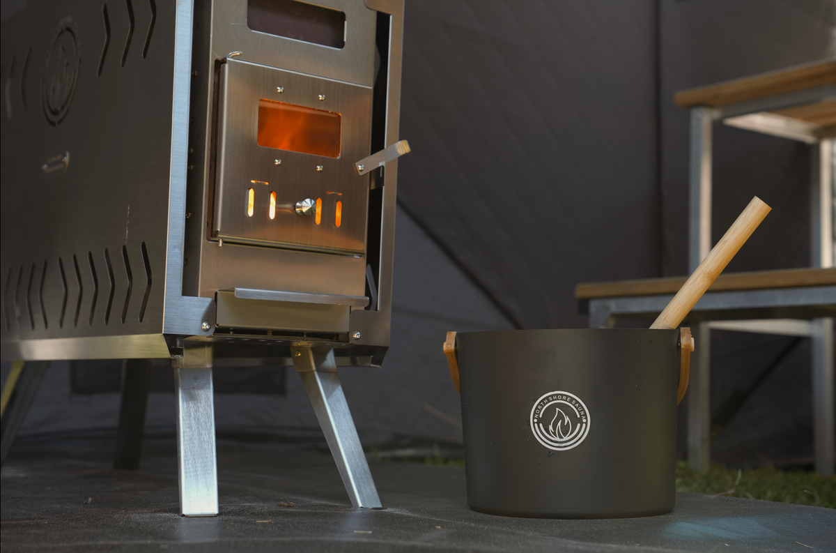 Black 5L aluminum sauna bucket and bamboo handled ladle set beside a lit wood burning stove inside a tent sauna