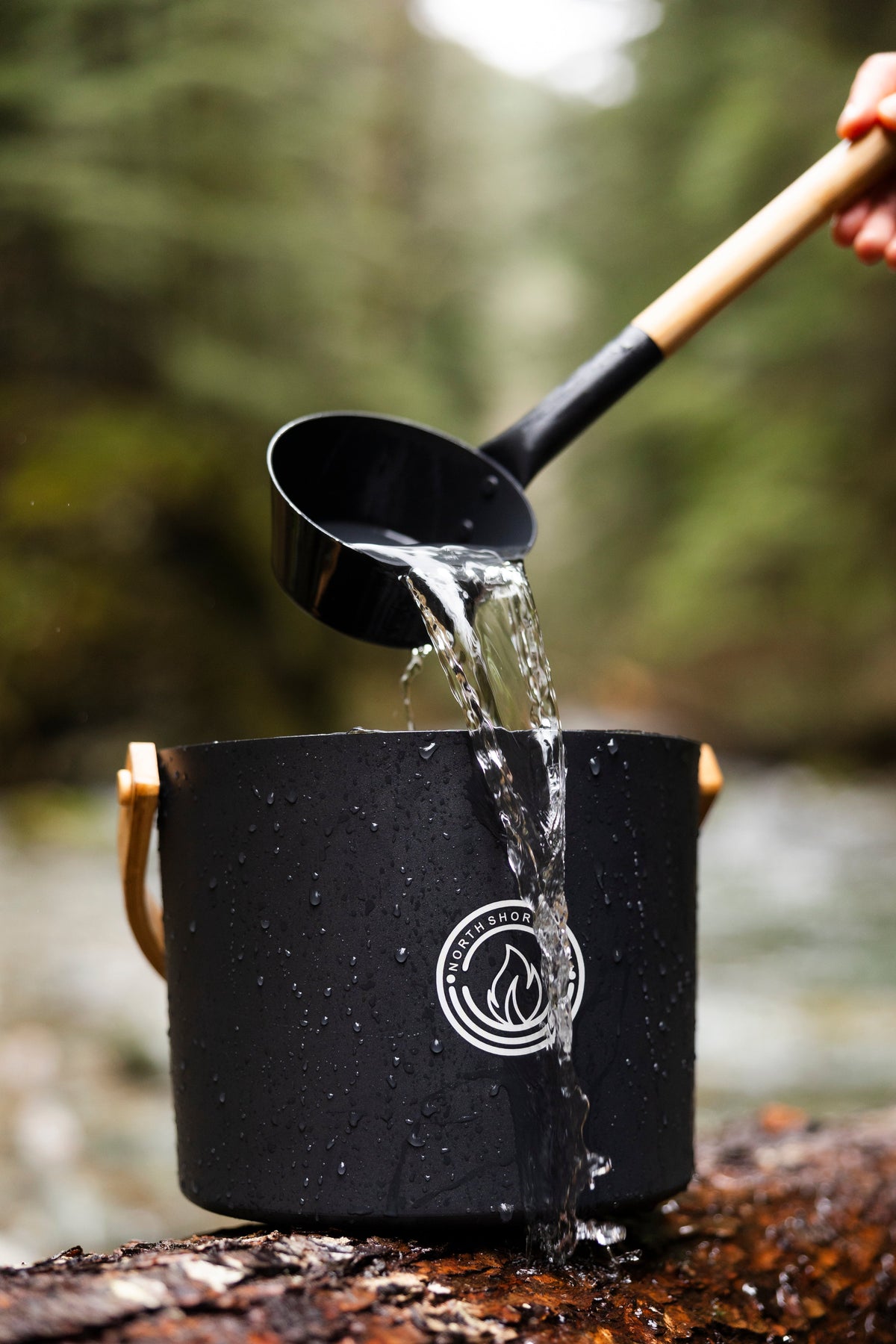 Black aluminum sauna bucket with bamboo handles and a bamboo handled ladle pouring water, shown outdoors beside a forest stream